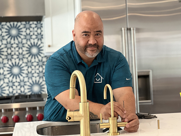 A Honey Homes handyman in turquoise polo shirt fixes a leak in a kitchen sink with brass fixtures