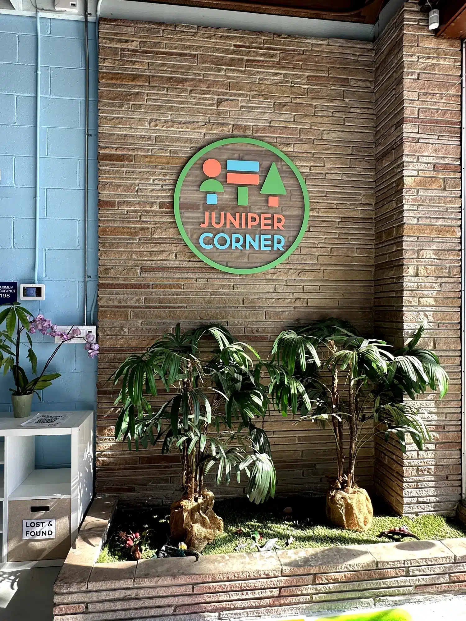Juniper Corner Business Sign Green-framed circular sign with colorful geometric shapes and orange and blue text on warm brown brick wall in Oakland, CA